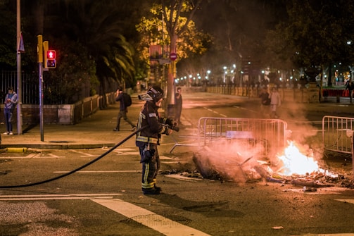 A firefighter in protective gear is standing on a street holding a hose, facing a small fire burning on the ground. The fire is surrounded by metal barricades, and it is nighttime with the scene illuminated by streetlights. Several people can be seen in the background, some walking and others standing, observing the situation.