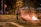 A firefighter in protective gear is standing on a street holding a hose, facing a small fire burning on the ground. The fire is surrounded by metal barricades, and it is nighttime with the scene illuminated by streetlights. Several people can be seen in the background, some walking and others standing, observing the situation.