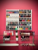 A neatly organized display of various nail polish bottles arranged on white racks against a pink wall. The polishes vary in color, including reds, blues, greens, yellows, and neutrals. Below the racks, two small potted plants sit on glass shelves, accompanied by additional nail polish bottles and what appears to be nail sample sticks.