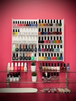 A neatly organized display of various nail polish bottles arranged on white racks against a pink wall. The polishes vary in color, including reds, blues, greens, yellows, and neutrals. Below the racks, two small potted plants sit on glass shelves, accompanied by additional nail polish bottles and what appears to be nail sample sticks.