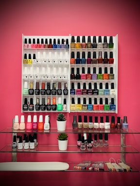 A neatly organized display of various nail polish bottles arranged on white racks against a pink wall. The polishes vary in color, including reds, blues, greens, yellows, and neutrals. Below the racks, two small potted plants sit on glass shelves, accompanied by additional nail polish bottles and what appears to be nail sample sticks.