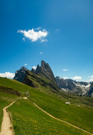 A picturesque mountain landscape with a clear blue sky, green grassy hills, and a rugged mountain peak in the background. A winding path leads through the grassy area, with a few hikers walking along it.