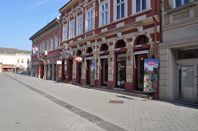A street scene features a row of historical buildings with ornate facades, arched windows, and decorative stonework. The street is empty, creating a serene and quiet atmosphere. There is a colorful signboard with the word 'COTTON' prominently displayed.