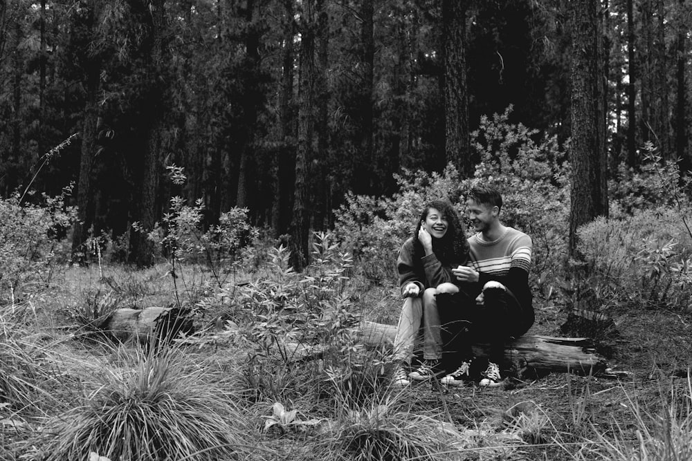 greyscale photo of woman and man sitting on log near forest