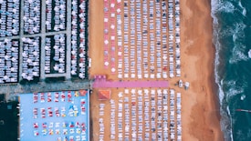An aerial view of a beach lined with numerous white sun loungers arranged in neat rows on sand. To the left of the image, a section shows a pool area with vibrant umbrellas and chairs. Waves from the ocean gently break on the shore on the right side.