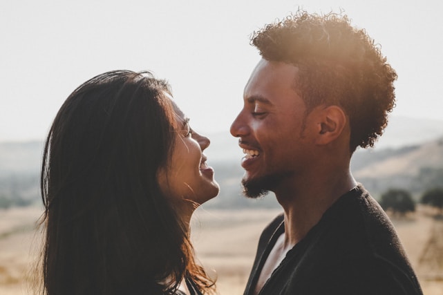 A joyful couple smiling warmly, standing close in a sunlit garden.