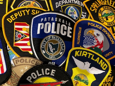 A collection of embroidered law enforcement patches from various departments, featuring badges with designs of eagles, flags, and department names. The colors are vibrant, and the patches are densely arranged, showcasing detailed stitching and a variety of insignias.