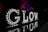 Illuminated letters spelling out 'GLOW' are displayed against a dark background. Each letter is adorned with bright lights, and the reflection of the letters can be seen on a shiny surface below. In the background, trees are illuminated with vibrant purple and red lights.
