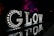Illuminated letters spelling out 'GLOW' are displayed against a dark background. Each letter is adorned with bright lights, and the reflection of the letters can be seen on a shiny surface below. In the background, trees are illuminated with vibrant purple and red lights.