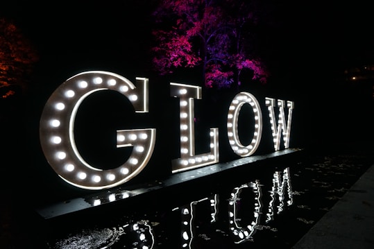 Illuminated letters spelling out 'GLOW' are displayed against a dark background. Each letter is adorned with bright lights, and the reflection of the letters can be seen on a shiny surface below. In the background, trees are illuminated with vibrant purple and red lights.