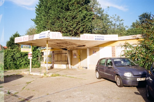 An abandoned car wash with faded and peeling signage under a canopy. Overgrown bushes surround the building, and an old, dusty car is parked in front of it. The scene is set in daylight with clear skies.