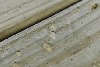 Water droplets scattered across a wooden surface, each droplet creating a small, clear reflection. The wood appears weathered with visible grain and slight discoloration.