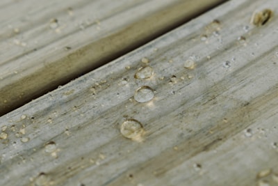 Water droplets scattered across a wooden surface, each droplet creating a small, clear reflection. The wood appears weathered with visible grain and slight discoloration.