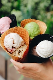 A collection of four ice cream cones held by a hand, featuring a variety of flavors and colors. The cones include waffle cones and dark cones with scoops of ice cream in pink, green, white, and brown tones.