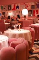A stylish dining area with plush pink chairs and white tablecloths, elegantly arranged with gold lamps on each table. The ambient lighting creates a warm and inviting atmosphere. The walls are adorned with modern art prints, adding a contemporary flair to the decor. Diners can be seen in the background, engaged in conversation.