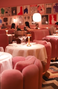 A stylish dining area with plush pink chairs and white tablecloths, elegantly arranged with gold lamps on each table. The ambient lighting creates a warm and inviting atmosphere. The walls are adorned with modern art prints, adding a contemporary flair to the decor. Diners can be seen in the background, engaged in conversation.