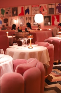 A stylish dining area with plush pink chairs and white tablecloths, elegantly arranged with gold lamps on each table. The ambient lighting creates a warm and inviting atmosphere. The walls are adorned with modern art prints, adding a contemporary flair to the decor. Diners can be seen in the background, engaged in conversation.