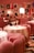 A stylish dining area with plush pink chairs and white tablecloths, elegantly arranged with gold lamps on each table. The ambient lighting creates a warm and inviting atmosphere. The walls are adorned with modern art prints, adding a contemporary flair to the decor. Diners can be seen in the background, engaged in conversation.