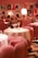 A stylish dining area with plush pink chairs and white tablecloths, elegantly arranged with gold lamps on each table. The ambient lighting creates a warm and inviting atmosphere. The walls are adorned with modern art prints, adding a contemporary flair to the decor. Diners can be seen in the background, engaged in conversation.