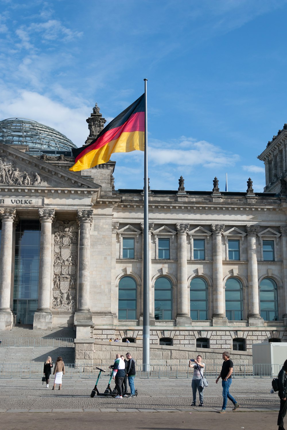 gente caminando al lado del edificio con la bandera ondeando de Alemania en el mástil