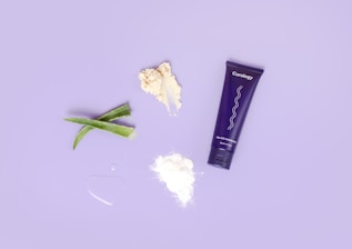 A flat lay composition features a purple tube labeled 'Curology' alongside various skincare ingredients such as aloe vera leaves, a scoop of white cream, a small pile of white powder, and a droplet of liquid. These items are arranged on a light purple background.