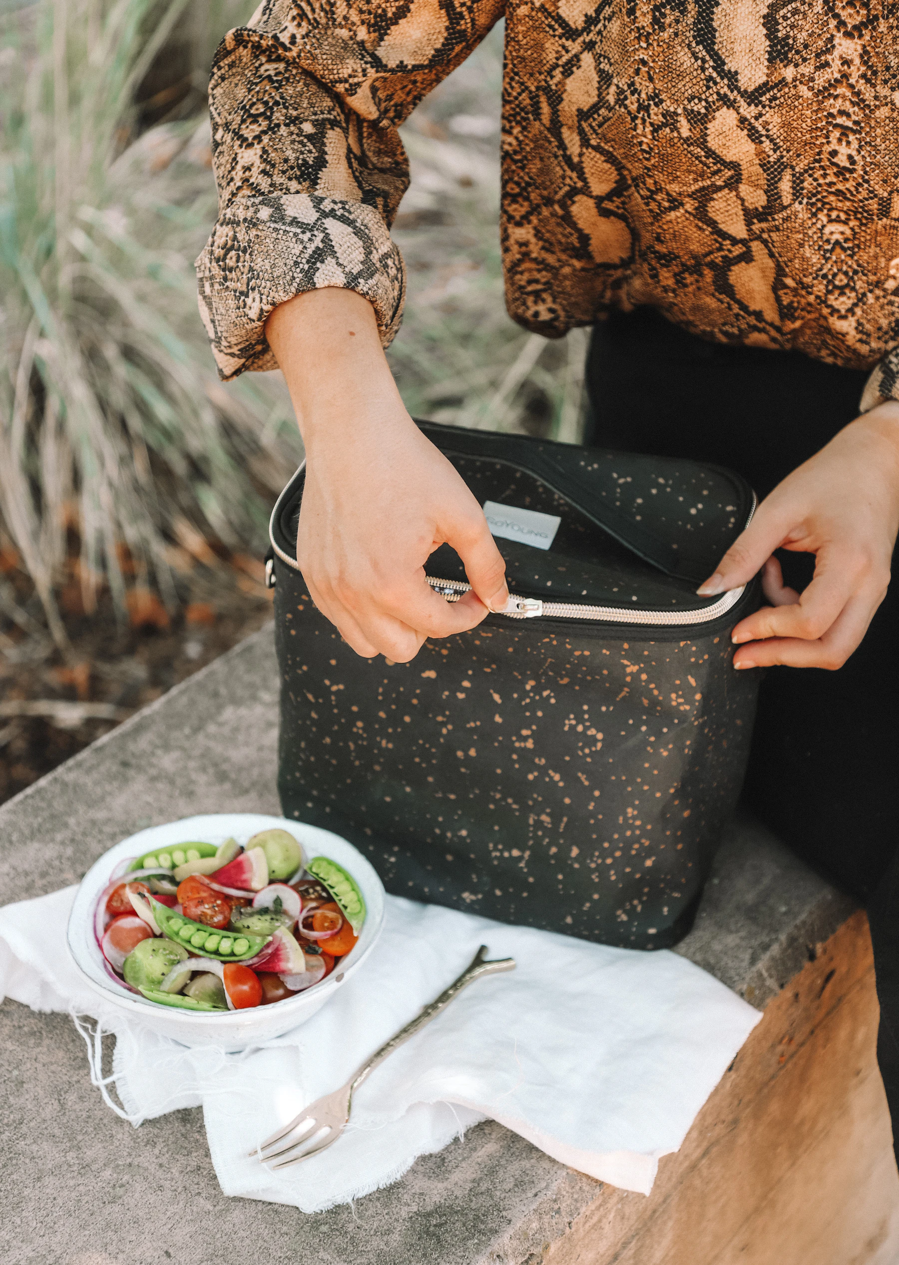 A woman zipping up a dark insulated lunch bag on a stone bench beside a bowl of salad