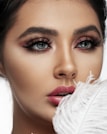 A close-up portrait of a person with striking blue eyes and dramatic makeup. The eyelashes are voluminous and accentuated with dark eyeliner and pink eyeshadow. The lips are full and covered with a vibrant pink lipstick. A white feather is held close to the lips, adding an element of softness.