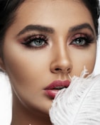 A close-up portrait of a person with striking blue eyes and dramatic makeup. The eyelashes are voluminous and accentuated with dark eyeliner and pink eyeshadow. The lips are full and covered with a vibrant pink lipstick. A white feather is held close to the lips, adding an element of softness.