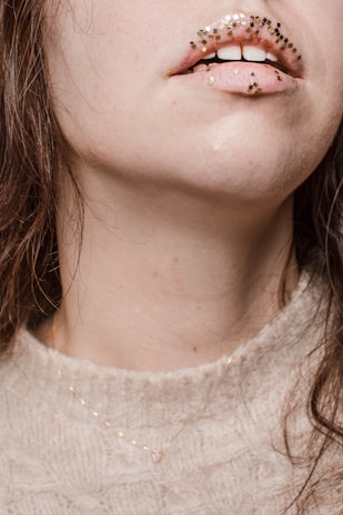 A close-up of a person's mouth and lower face, adorned with small, square glitter pieces in shades of gold and black on the lips. The person has fair skin, and their hair is dark brown. They are wearing a light beige knitted sweater and a delicate gold necklace.
