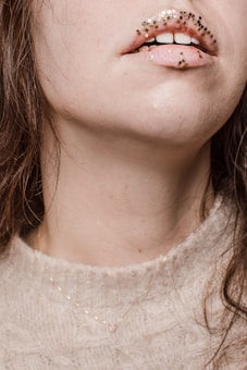 A close-up of a person's mouth and lower face, adorned with small, square glitter pieces in shades of gold and black on the lips. The person has fair skin, and their hair is dark brown. They are wearing a light beige knitted sweater and a delicate gold necklace.
