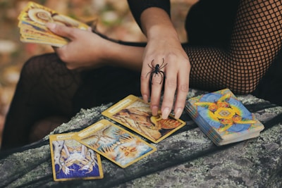 A person dressed in a black fishnet outfit holds a set of tarot cards. The hand is adorned with a large spider ring, and a deck of cards lies nearby on a textured surface. The scene is set outdoors with autumn leaves visible in the background.