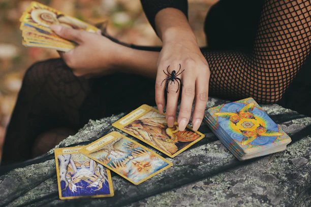A person dressed in a black fishnet outfit holds a set of tarot cards. The hand is adorned with a large spider ring, and a deck of cards lies nearby on a textured surface. The scene is set outdoors with autumn leaves visible in the background.