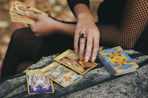 A person dressed in a black fishnet outfit holds a set of tarot cards. The hand is adorned with a large spider ring, and a deck of cards lies nearby on a textured surface. The scene is set outdoors with autumn leaves visible in the background.