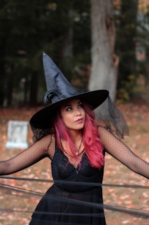 A woman with pink hair, wearing a black witch's hat and a black lace dress with mesh sleeves, is standing outdoors. She is in front of a background of trees with autumn leaves on the ground.