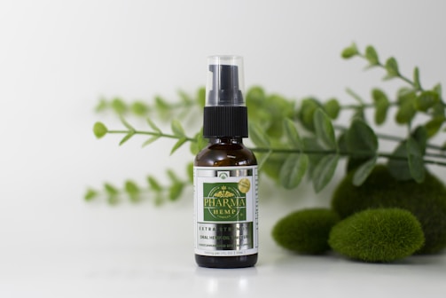 A small glass bottle with a spray nozzle labeled 'Pharma Hemp' is placed in front of a backdrop of green leaves and mossy stones on a white surface.