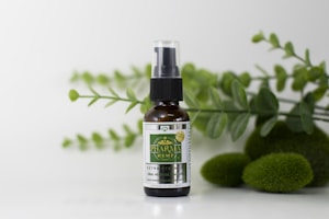 A small glass bottle with a spray nozzle labeled 'Pharma Hemp' is placed in front of a backdrop of green leaves and mossy stones on a white surface.