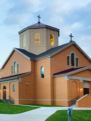 A brown brick church building with arched windows, a central octagonal dome with a cross, and another cross at the peak of the roof. The structure is set against a backdrop of a cloudy sky, and a pathway leads up to the entrance, flanked by grass.