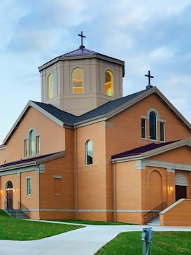 A brown brick church building with arched windows, a central octagonal dome with a cross, and another cross at the peak of the roof. The structure is set against a backdrop of a cloudy sky, and a pathway leads up to the entrance, flanked by grass.
