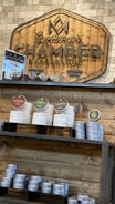 A wooden sign with the logo and name 'Gentlemen's Chamber Barbershop' hangs on a stone wall. Below it, shelves hold stacked tins and jars of shaving products in various colors, including red, green, and brown. The display has a rustic and organized appearance.