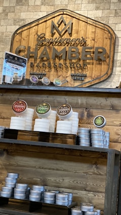 A wooden sign with the logo and name 'Gentlemen's Chamber Barbershop' hangs on a stone wall. Below it, shelves hold stacked tins and jars of shaving products in various colors, including red, green, and brown. The display has a rustic and organized appearance.