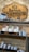 A wooden sign with the logo and name 'Gentlemen's Chamber Barbershop' hangs on a stone wall. Below it, shelves hold stacked tins and jars of shaving products in various colors, including red, green, and brown. The display has a rustic and organized appearance.
