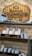 A wooden sign with the logo and name 'Gentlemen's Chamber Barbershop' hangs on a stone wall. Below it, shelves hold stacked tins and jars of shaving products in various colors, including red, green, and brown. The display has a rustic and organized appearance.
