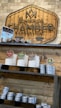 A wooden sign with the logo and name 'Gentlemen's Chamber Barbershop' hangs on a stone wall. Below it, shelves hold stacked tins and jars of shaving products in various colors, including red, green, and brown. The display has a rustic and organized appearance.