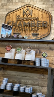 A wooden sign with the logo and name 'Gentlemen's Chamber Barbershop' hangs on a stone wall. Below it, shelves hold stacked tins and jars of shaving products in various colors, including red, green, and brown. The display has a rustic and organized appearance.