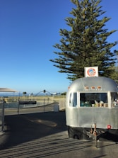 A shiny silver food trailer is parked on a pavement, with a large tree and a clear blue sky in the background. The trailer has a small 'OPEN' sign in the window, and a sign with a logo on the roof. In the distance, there are some park structures and seating areas, surrounded by a fence, with a hint of the sea visible.