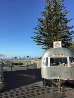 A shiny silver food trailer is parked on a pavement, with a large tree and a clear blue sky in the background. The trailer has a small 'OPEN' sign in the window, and a sign with a logo on the roof. In the distance, there are some park structures and seating areas, surrounded by a fence, with a hint of the sea visible.