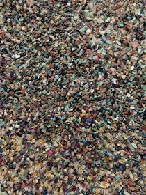 A variety of small, colorful pebbles and stones scattered across a surface, creating a textured and vibrant arrangement. The stones come in shades of red, green, blue, purple, and beige, among others, with a glossy finish that reflects light subtly.