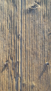 Close-up view of a wooden surface with distinct vertical grooves and textured patterns. The wood shows natural signs of wear, including knots and weathering marks.
