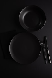 A minimalistic flat lay setup featuring a dark-colored dinner plate and bowl. Positioned on a black surface, the arrangement includes a knife and fork to the right of the plate. The overall composition emphasizes simplicity and elegance, with a focus on clear lines and shapes.
