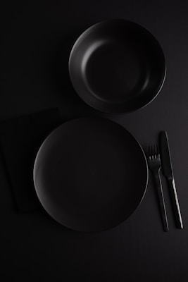 A minimalistic flat lay setup featuring a dark-colored dinner plate and bowl. Positioned on a black surface, the arrangement includes a knife and fork to the right of the plate. The overall composition emphasizes simplicity and elegance, with a focus on clear lines and shapes.
